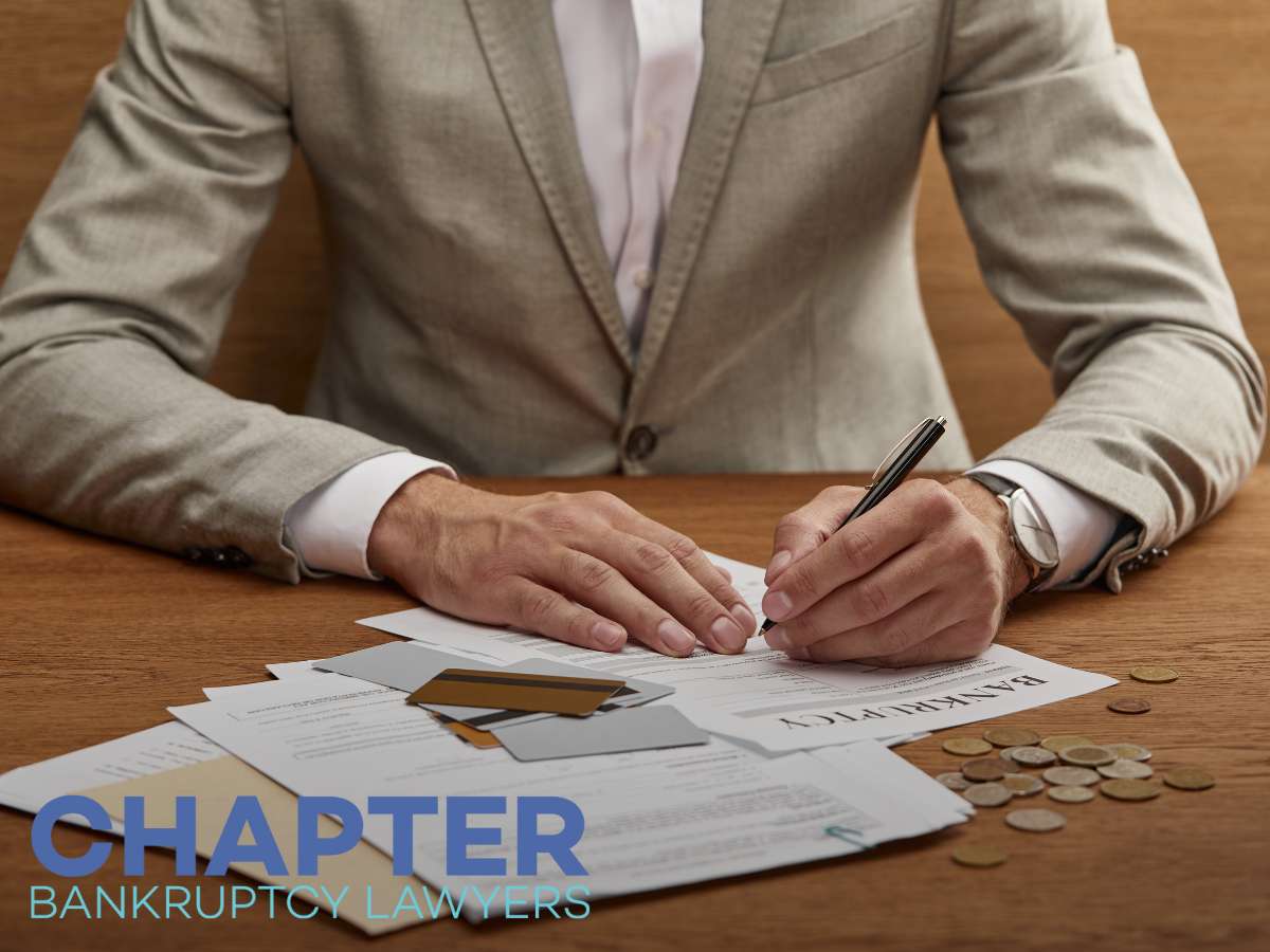 Person in a suit at a wooden desk, writing on bankruptcy documents surrounded by scattered coins and paperwork